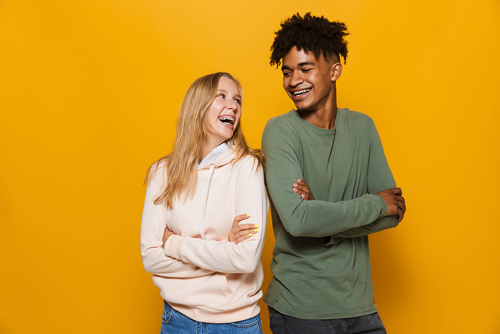 Two young adults laughing against a yellow background, highlighting the benefits of traditional braces.