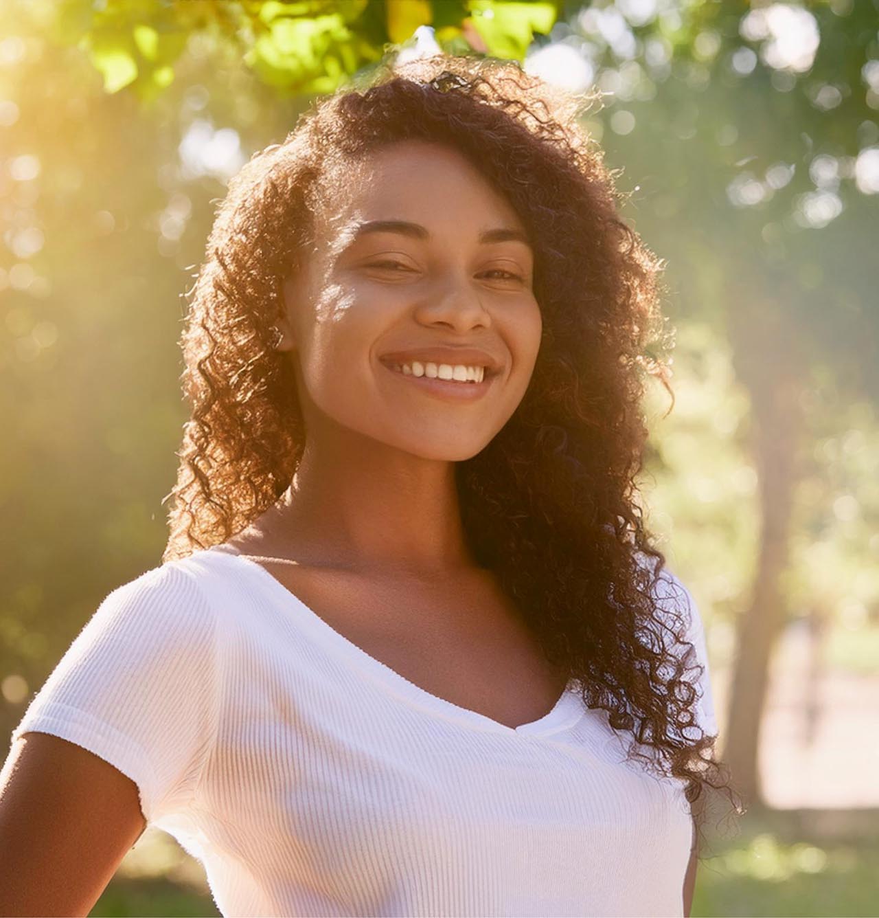 Smiling Black woman with curly hair, showing bright white teeth in an outdoor setting.